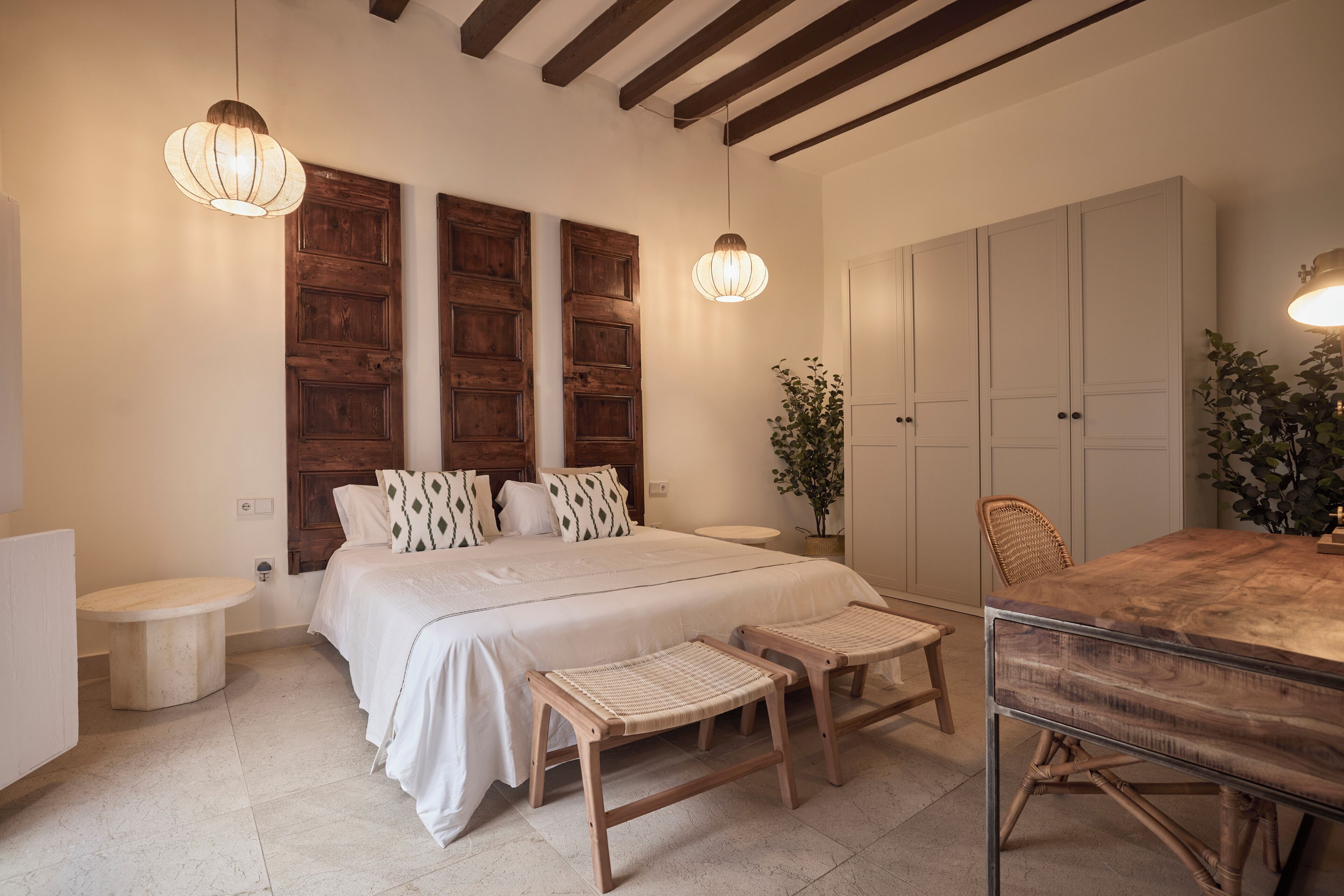 A bedroom with a large bed, two bedside tables, a wooden bench, a dining table, and a wardrobe.