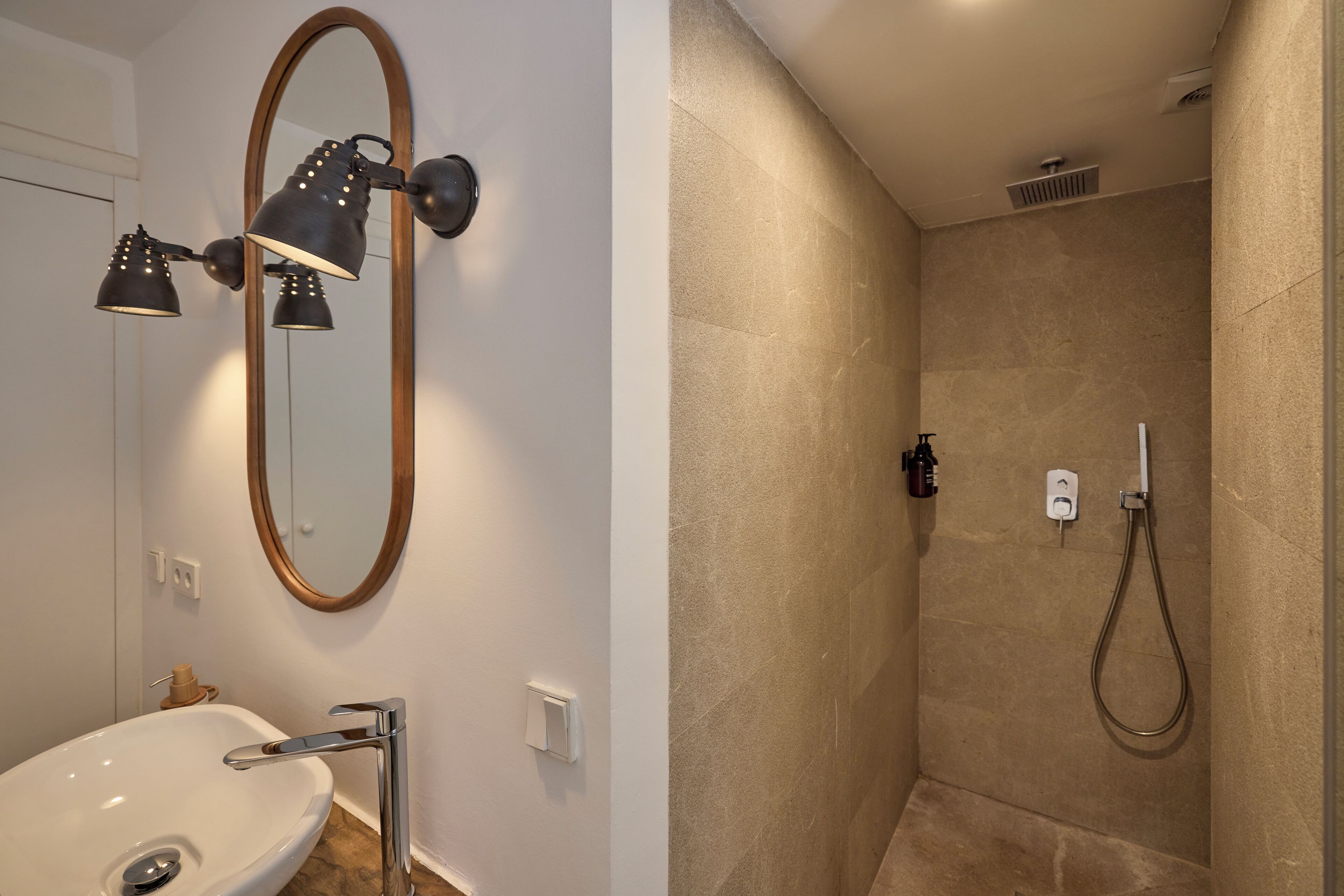 Een badkamer met een wit wastafel, een spiegel met houten lijst en een douche met een beige wand en een handdouche.