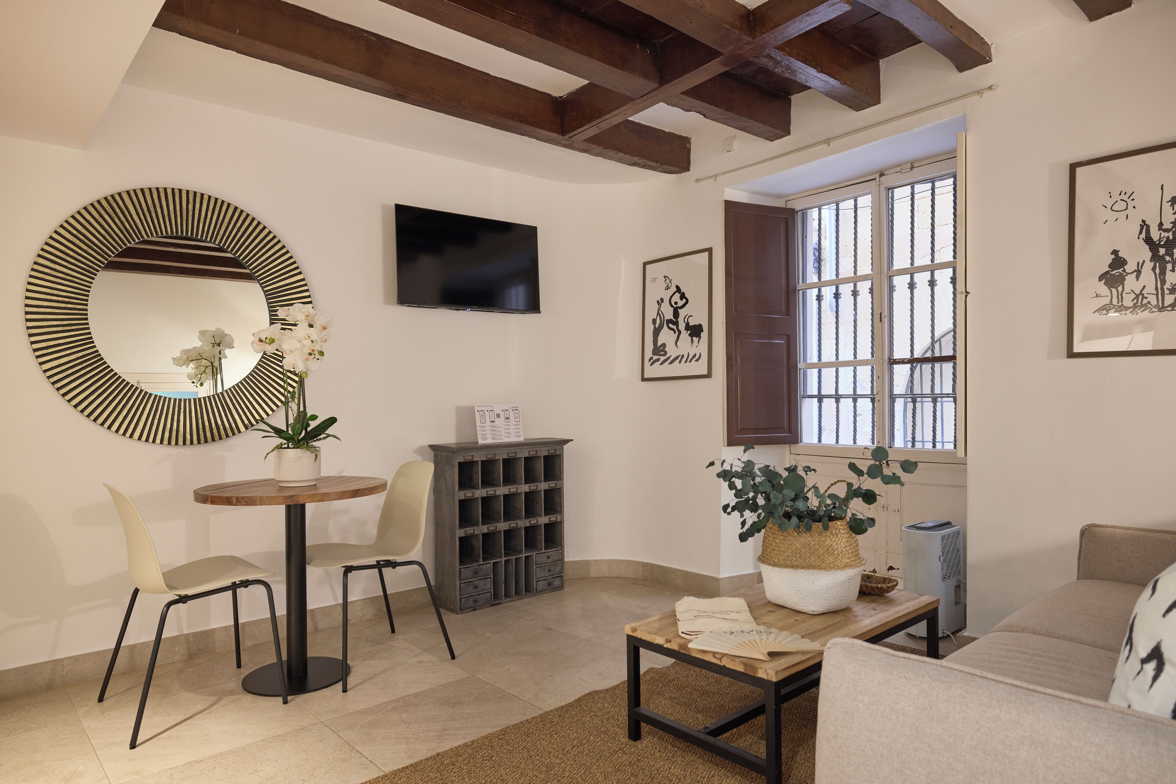 A living room with a round mirror, a television, a bookshelf, a sofa, and a window with a view.