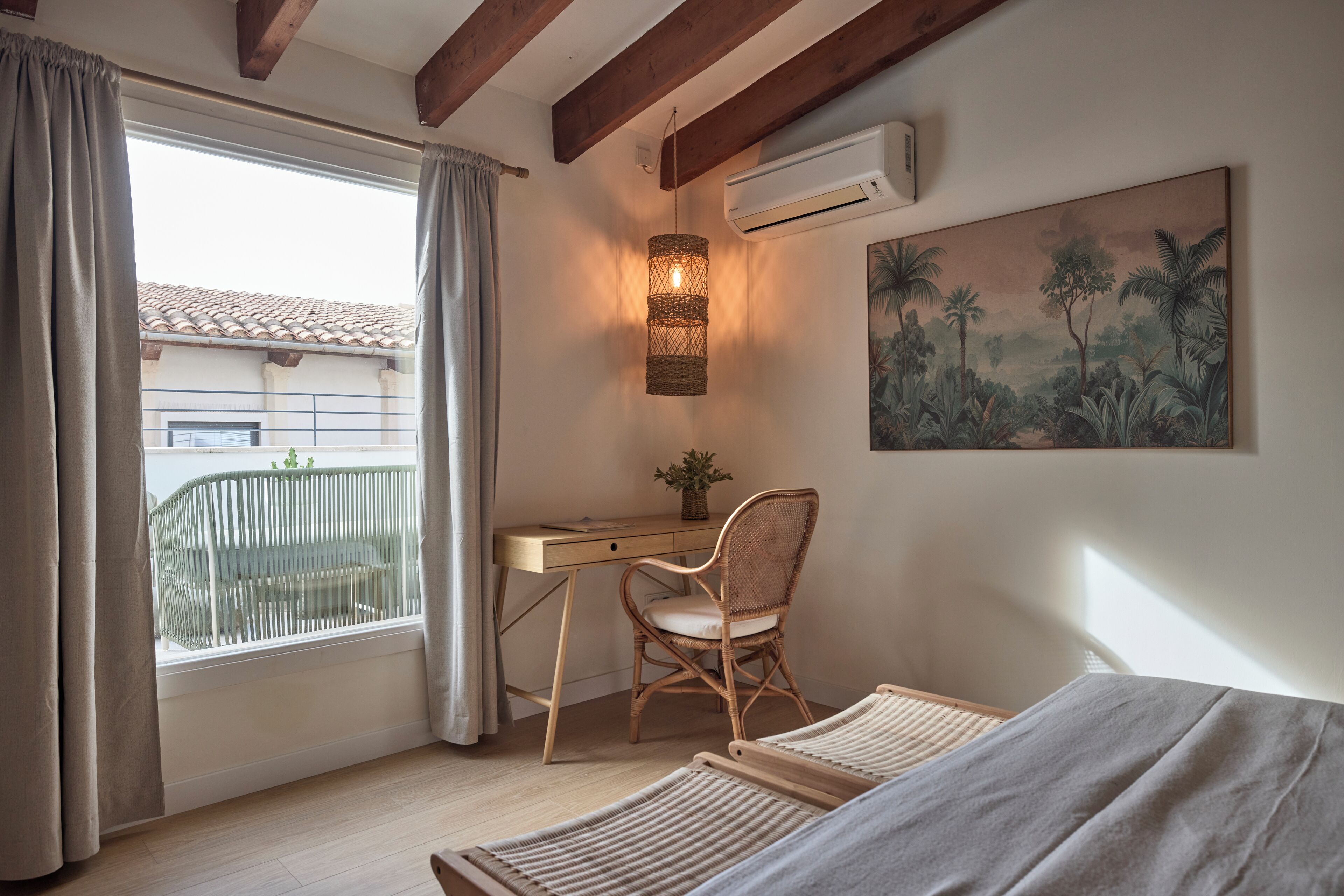 Un dormitorio con escritorio, una silla de mimbre, una cama y un cuadro de un paisaje tropical.