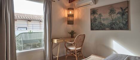 Un dormitorio con escritorio, una silla de mimbre, una cama y un cuadro de un paisaje tropical.