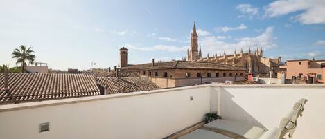 Una terraza en la azotea con sillones blancos, una palmera y vistas a edificios históricos con una torre destacada.