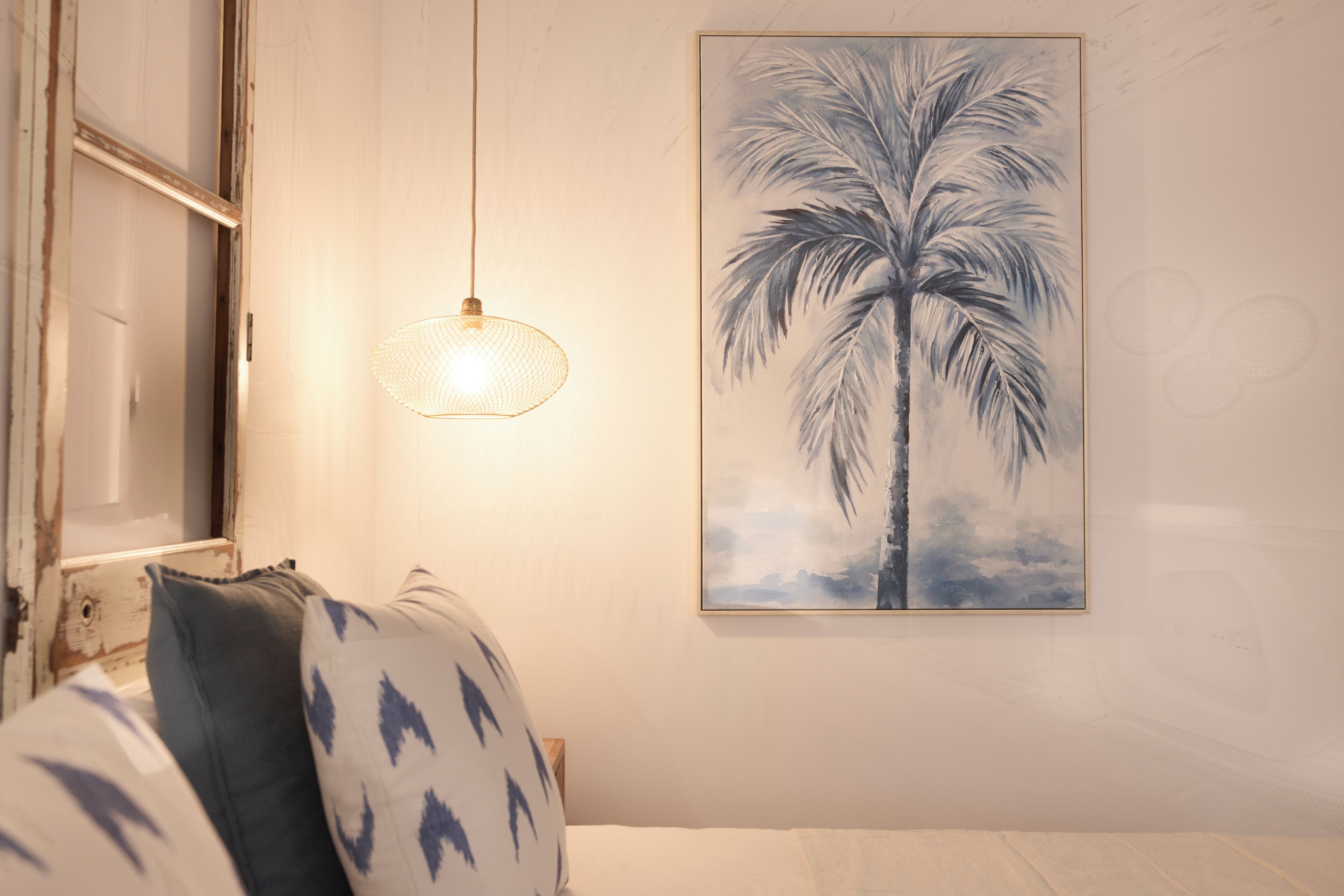A bedroom with a bed, pillows, a hanging light fixture, and a wall-mounted painting of a palm tree.