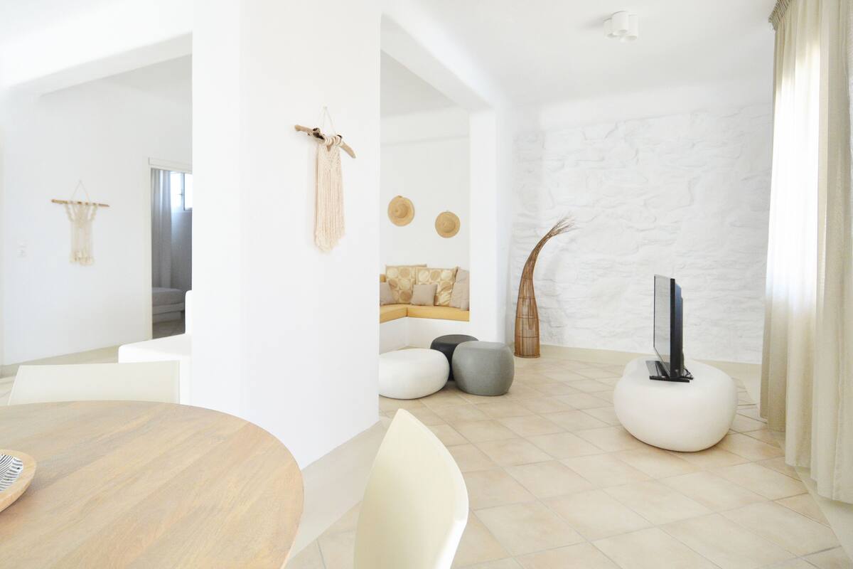 Una sala moderna con una mesa redonda de madera, sillas blancas, un sofá con cojines estampados, un televisor sobre un reposapiés blanco y una pared de piedra.