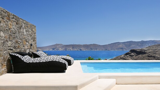 Área de piscina com muro de pedra e vista para o mar e montanhas.