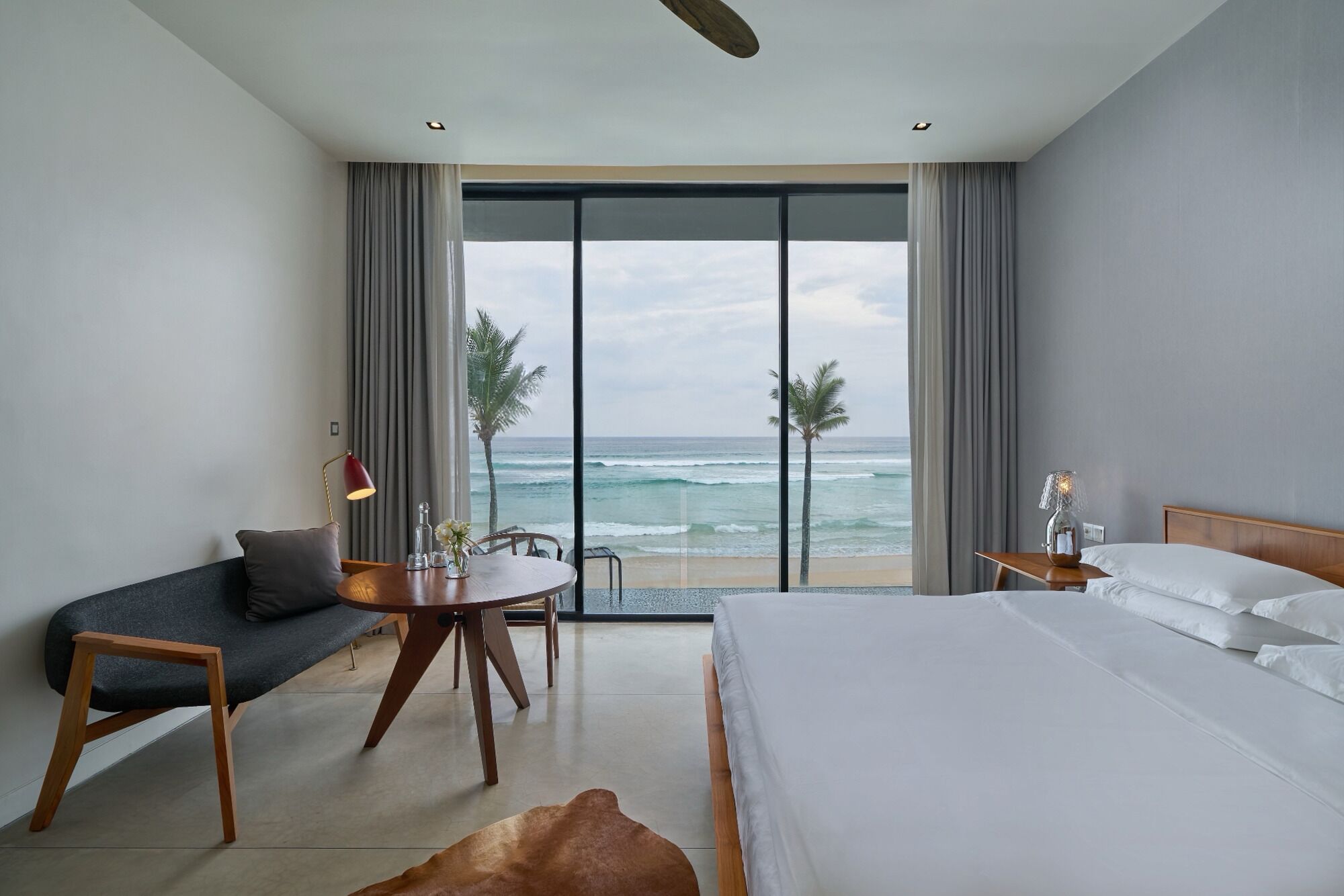 A hotel room with a bed, a desk, a chair, a small table with a lamp, and a view of the ocean.