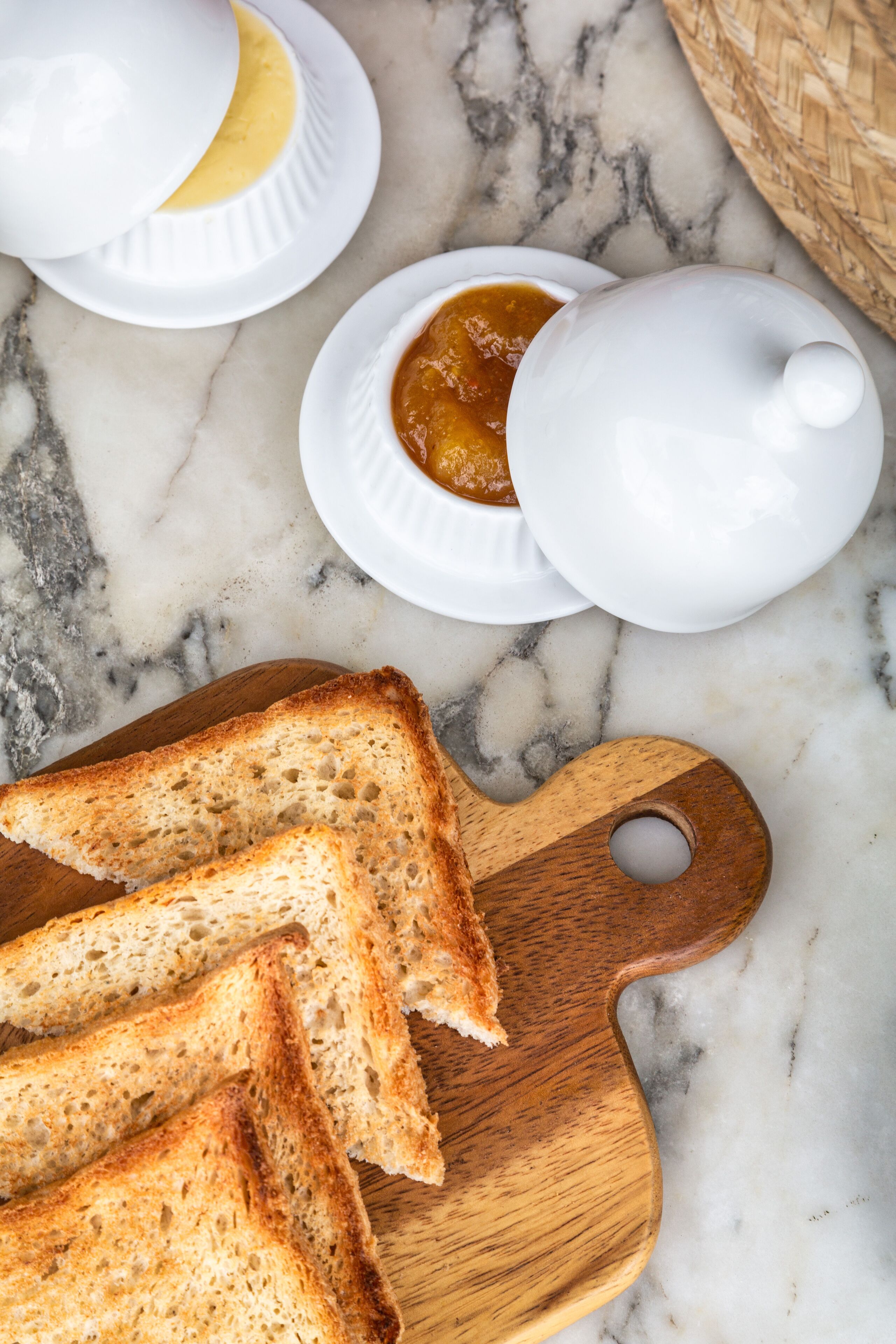 Un plan de travail en marbre sur lequel repose une planche à découper en bois, avec des tranches de pain grillé, une théière blanche et un petit bol de confiture.