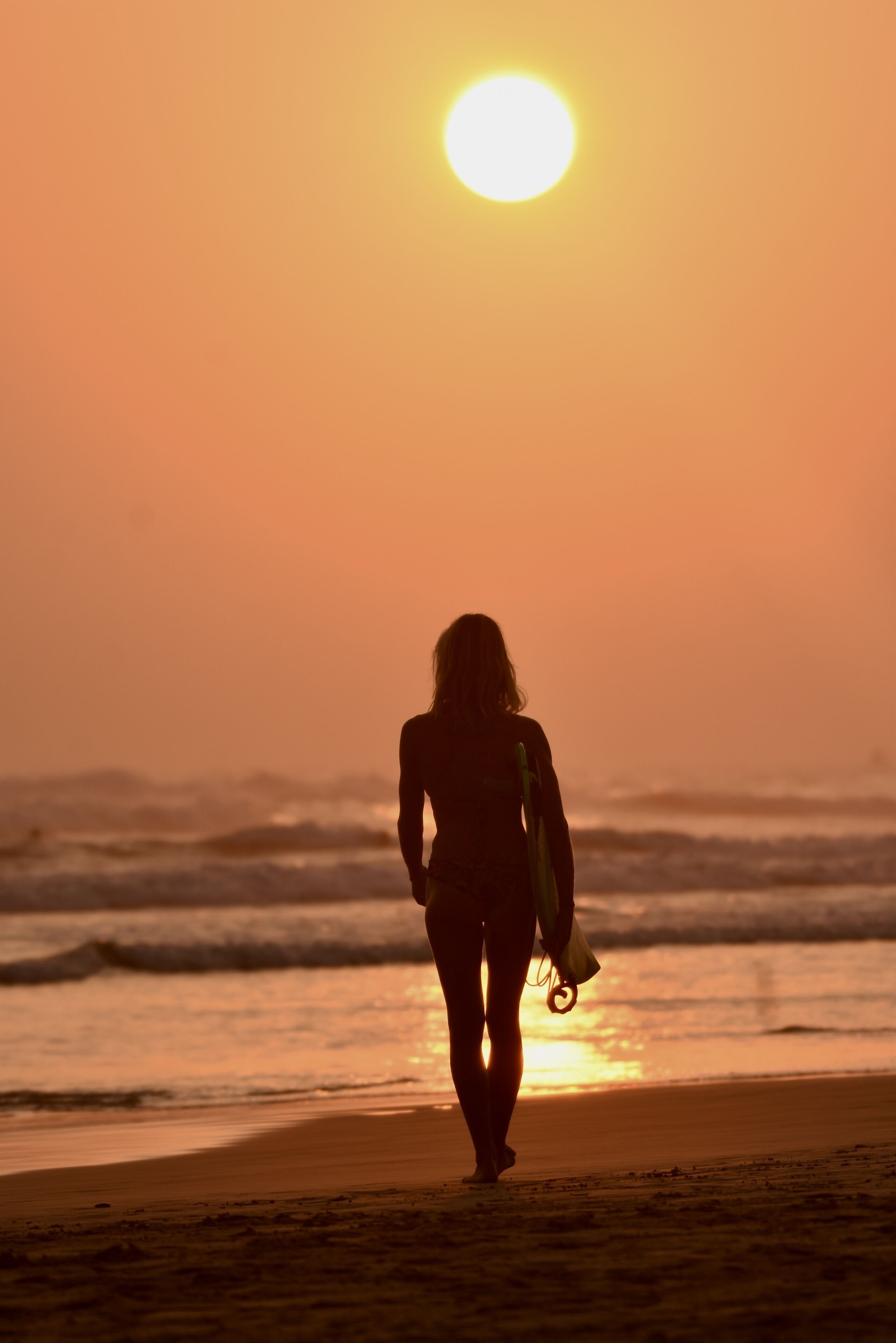 Uma pessoa caminhando na praia com uma prancha de surf, indo em direção ao oceano ao pôr do sol.