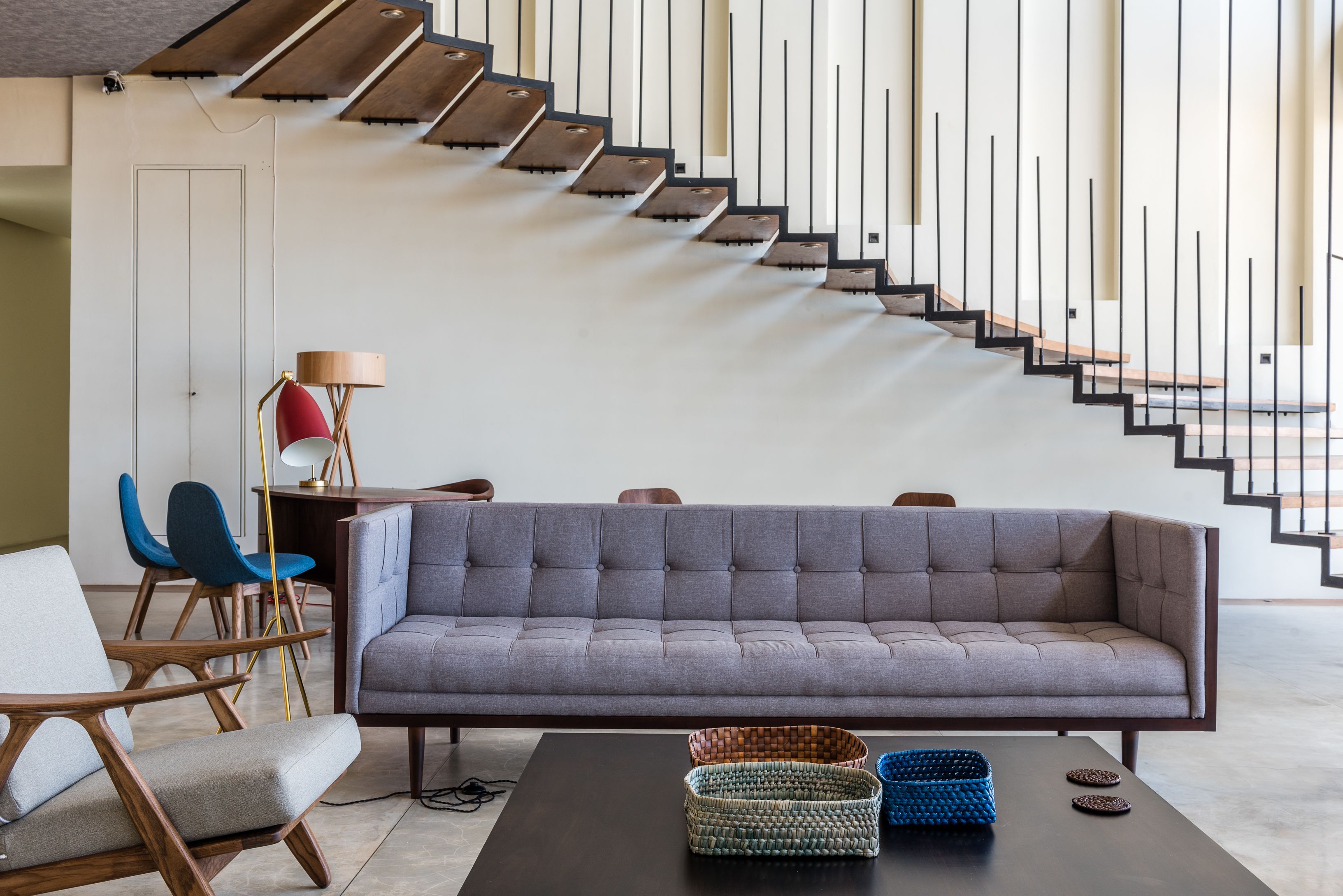 A modern living room with a grey sofa, wooden chairs, a black coffee table, and a staircase with black railings.