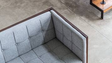 Un sofá tapizado gris con estructura de madera, ubicado sobre un suelo de hormigón con una escalera de madera al fondo.