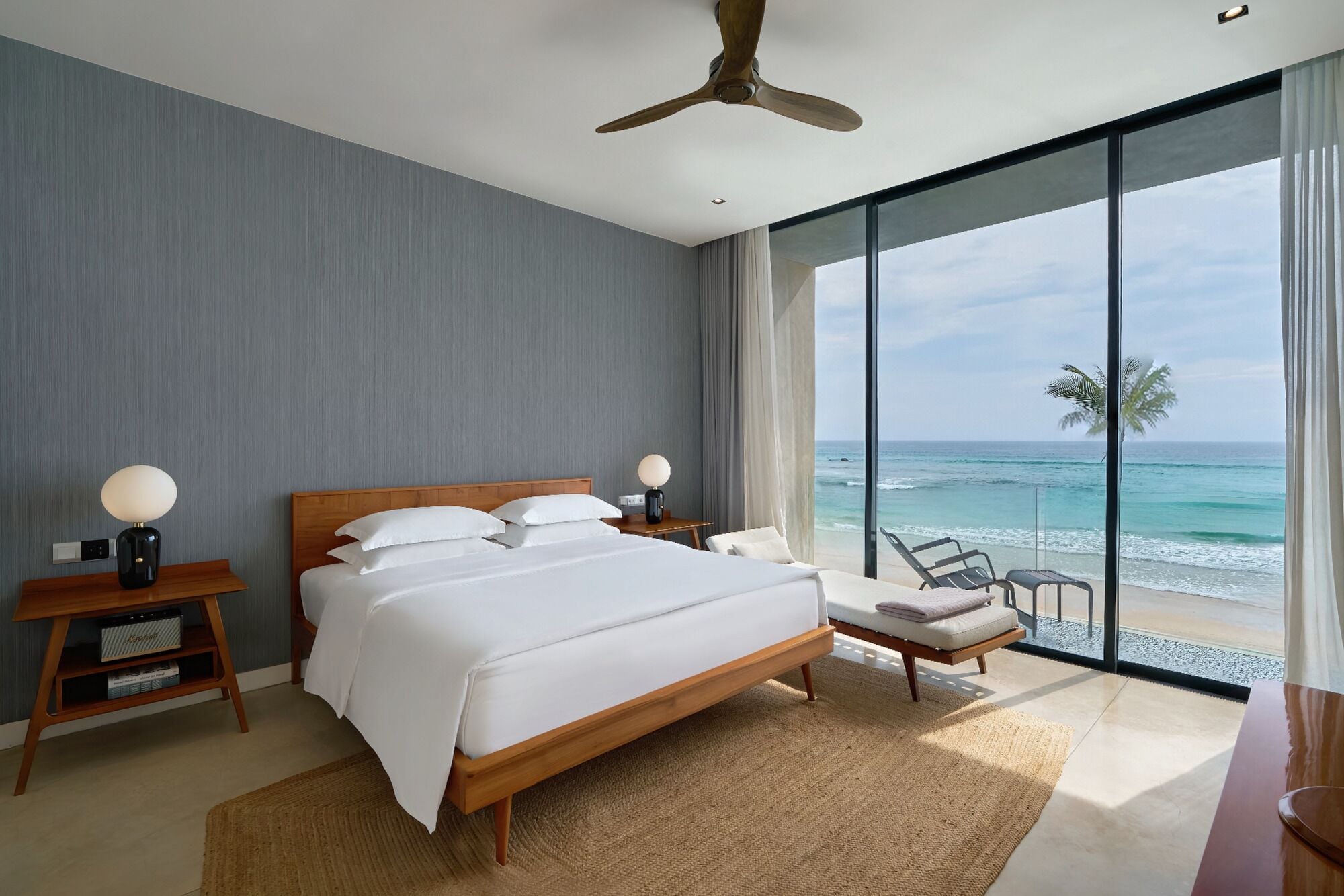 A modern hotel room with a large bed, a wooden nightstand, a desk, and a view of the ocean.