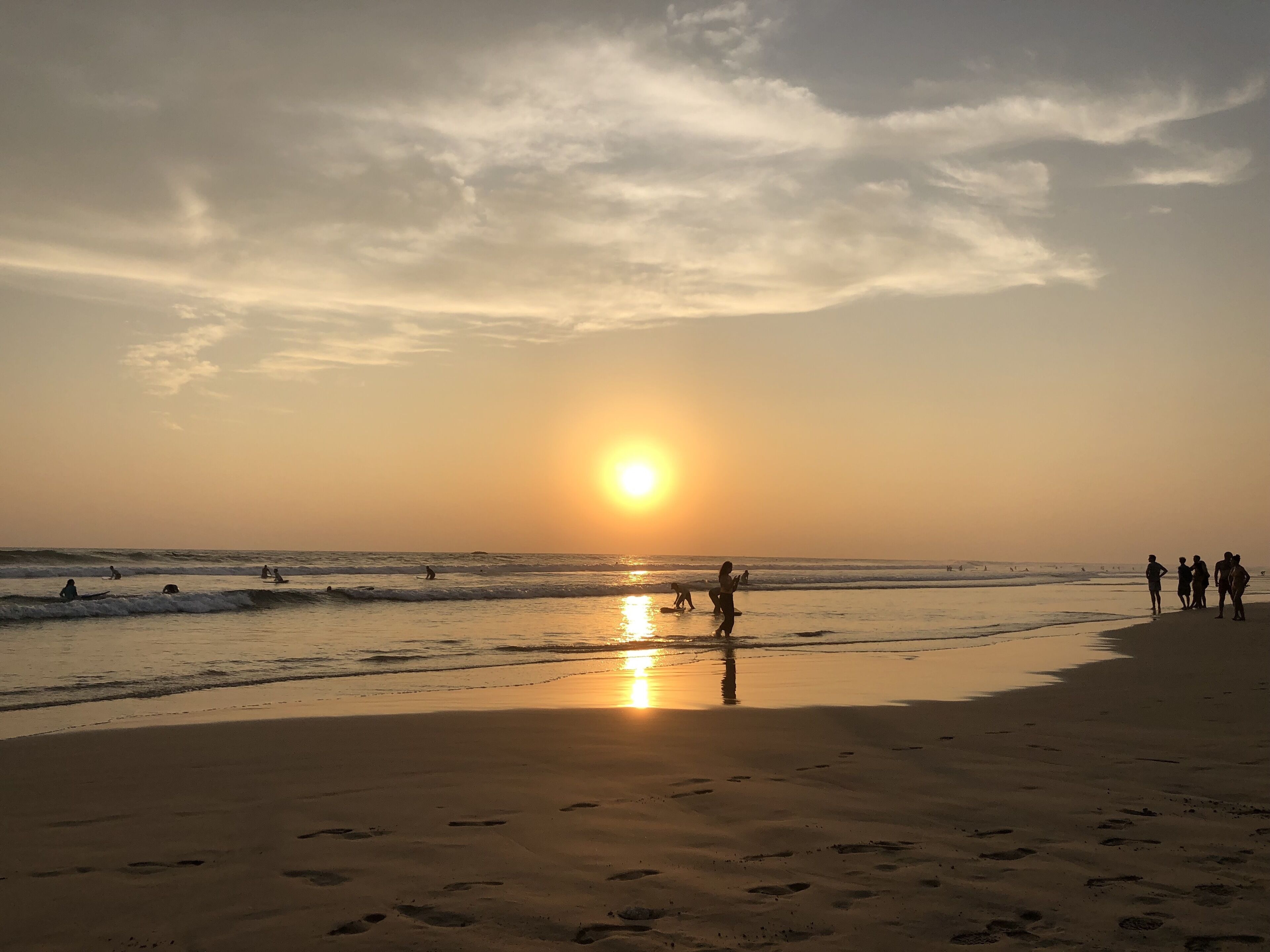 Een strandscène met een ondergaande zon aan de horizon, mensen die langs de kust wandelen en voetafdrukken in het zand.