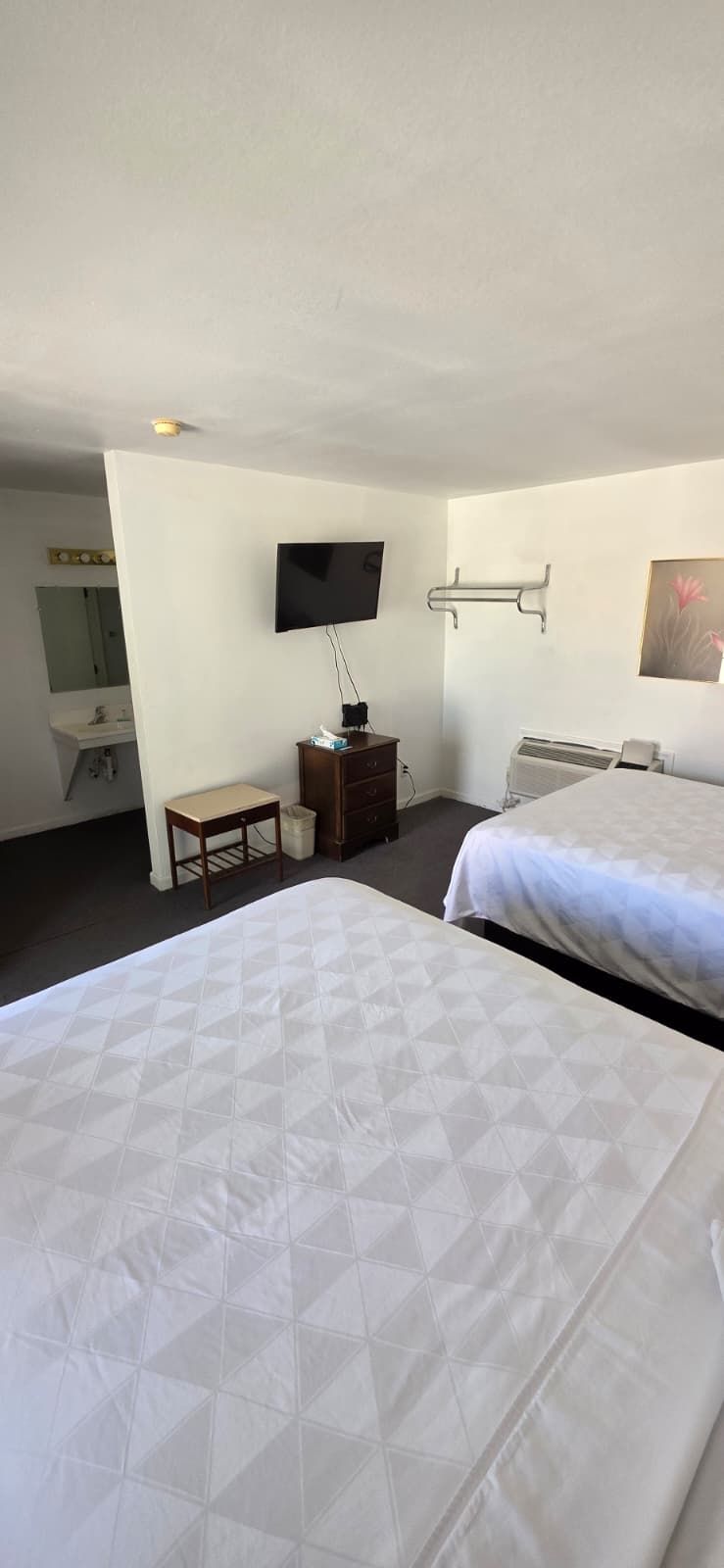 Habitación de hotel con dos camas, un televisor montado en la pared, una mesita pequeña y un baño visible a través de una puerta abierta.