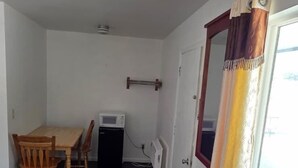 A narrow room with a dining table and chairs, a small black heater, a window with a curtain, and a metal rack.