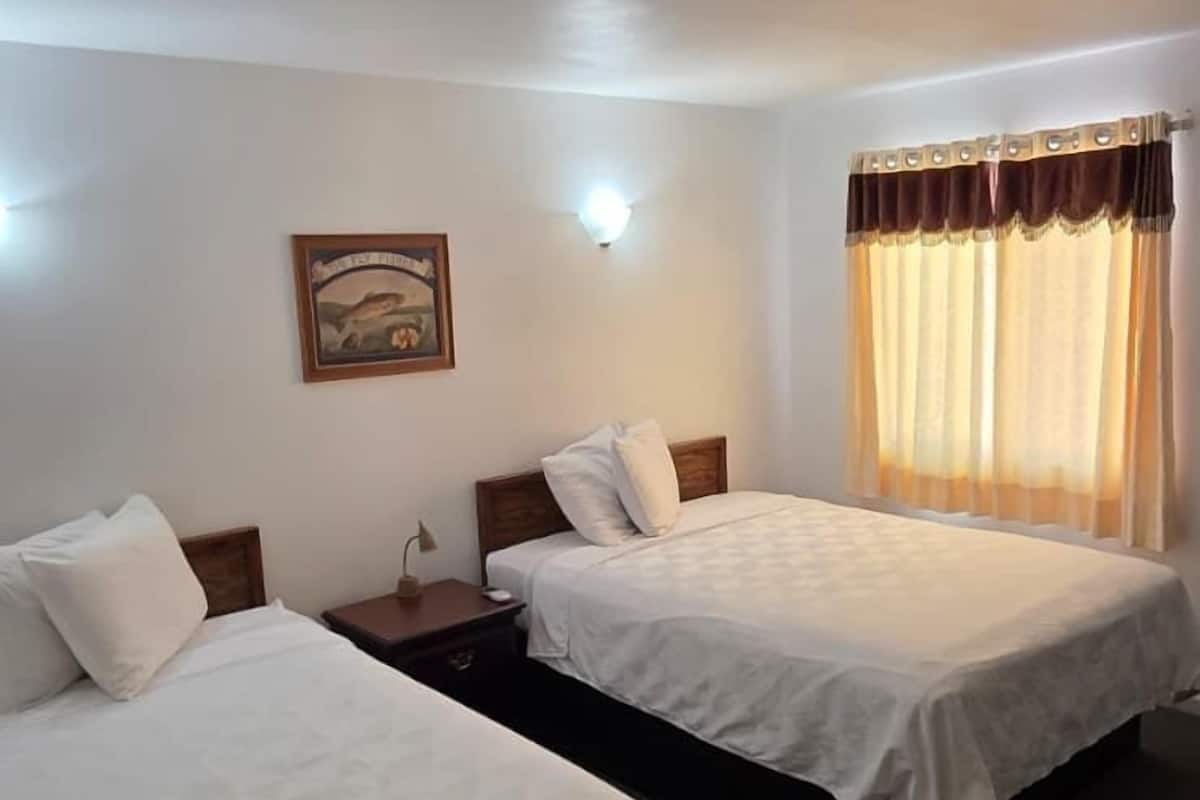 A hotel room with two single beds, a nightstand with a lamp, a framed picture on the wall, and a window with curtains.