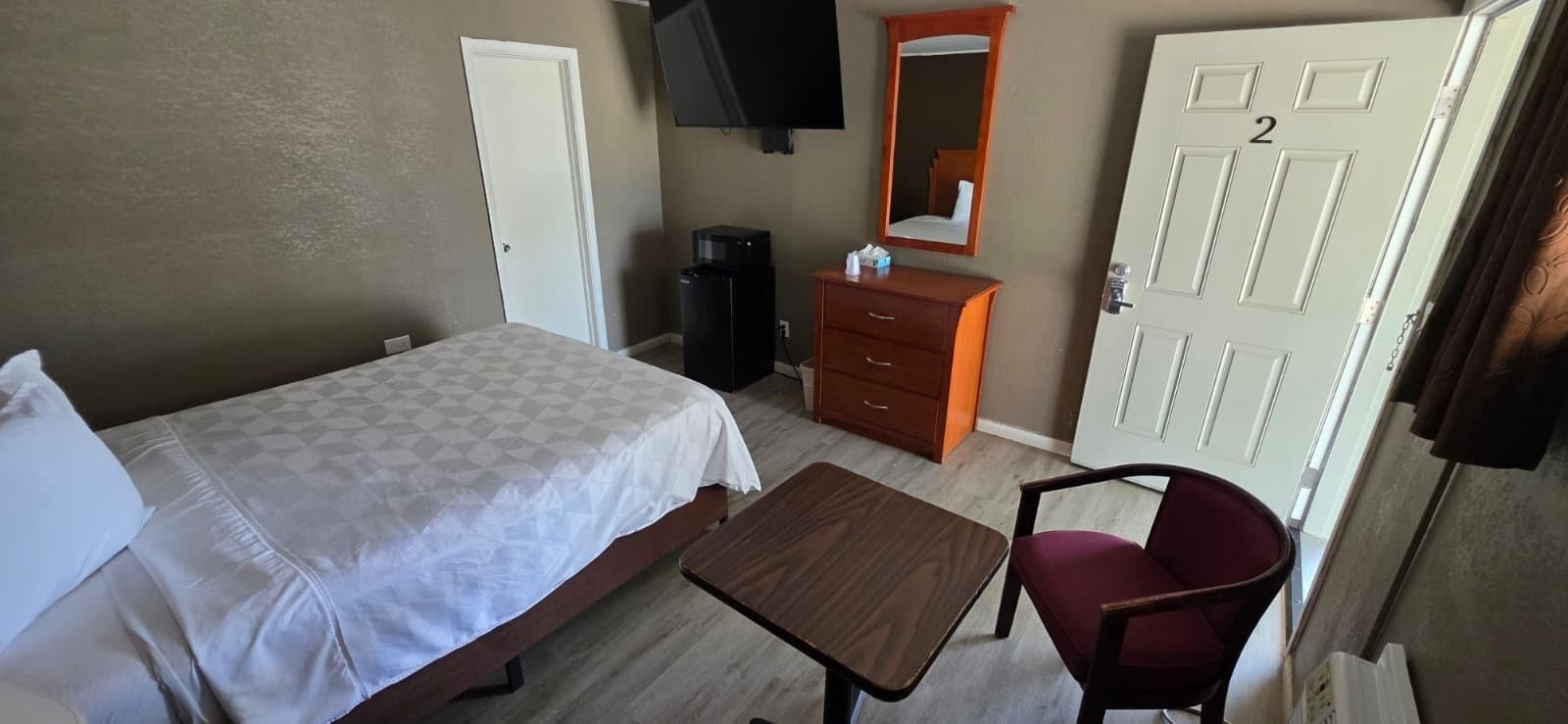 A hotel room with a bed, a small table, a chair, a dresser, a mirror, a TV, and a door with the number 2.