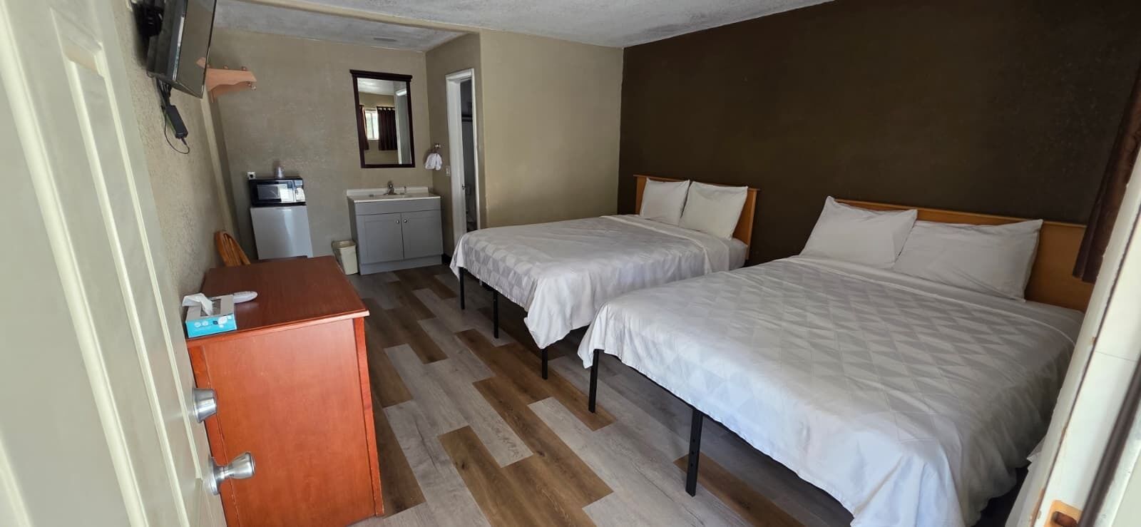 Habitación de hotel con dos camas, un escritorio de madera, una pequeña cocina y un espejo.