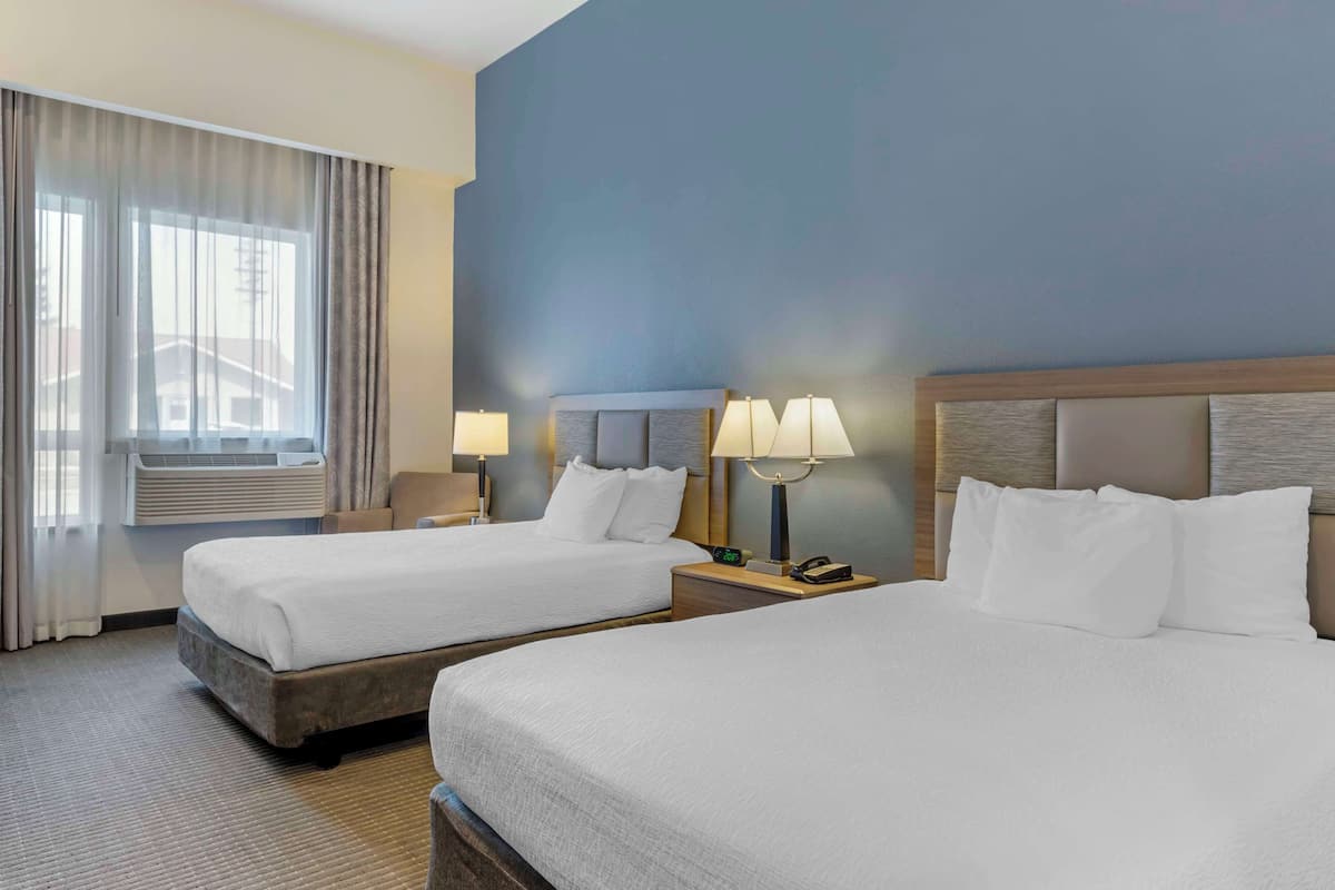A hotel room with two beds, a large window with curtains, an air conditioning unit, and bedside lamps.