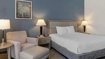 A hotel room with a large bed, a chair, a small table, two bedside lamps, a framed artwork on the wall, and a nightstand with a phone.