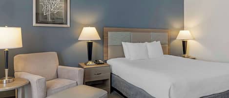 Habitación de hotel con una cama grande, una silla, una mesita, dos lámparas de noche, un cuadro enmarcado en la pared y una mesita de noche con un teléfono.