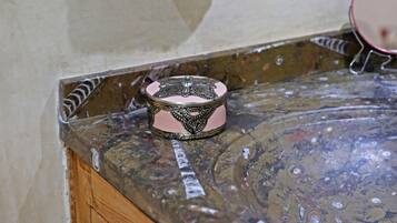 Un baño con un estante de mimbre que sostiene un recipiente rosa, un cuenco decorativo en la encimera y un espejo en la pared.