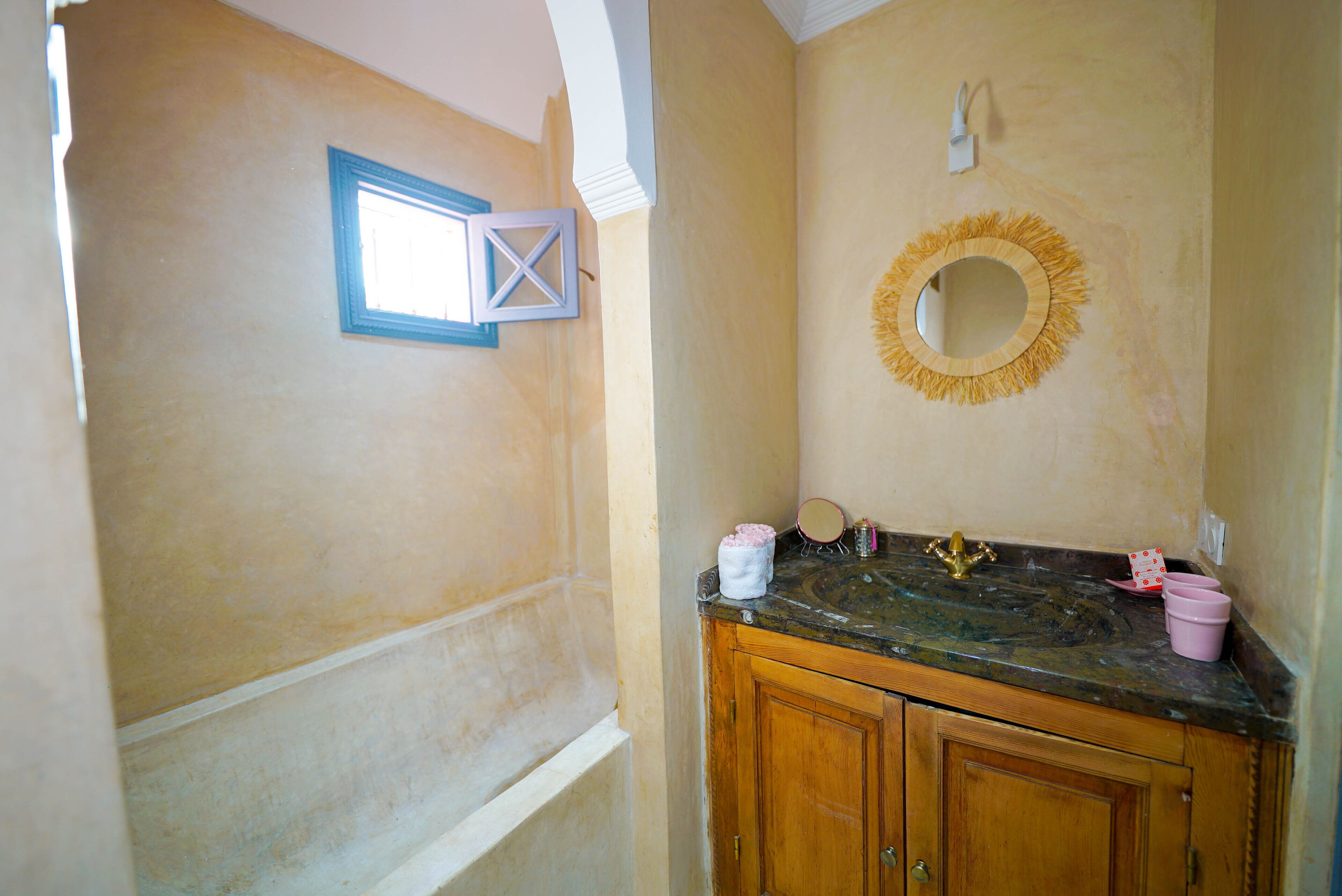 A bathroom with a wooden vanity, a sink with a marble countertop, a mirror with a decorative frame, and a small window with a blue frame.