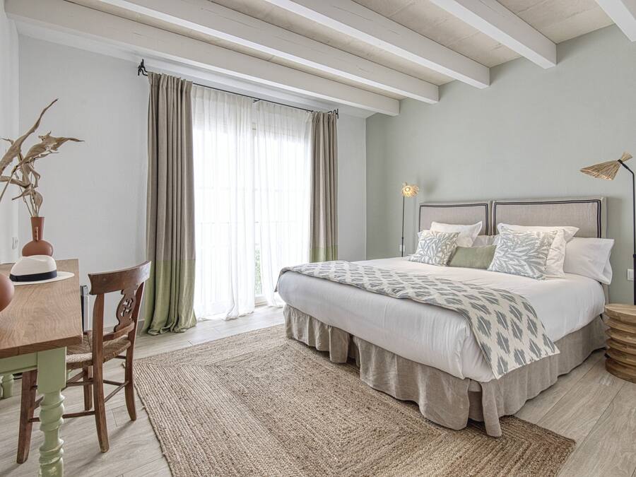 A bedroom with a bed, a desk, and a chair. There is a window with curtains, a lamp, and a rug.