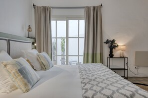 Ein ordentlich bezogenes Bett mit gemusterten Betttextilien, ein Nachttisch mit Lampe und Blick ins Freie durch ein Fenster mit Vorhängen.