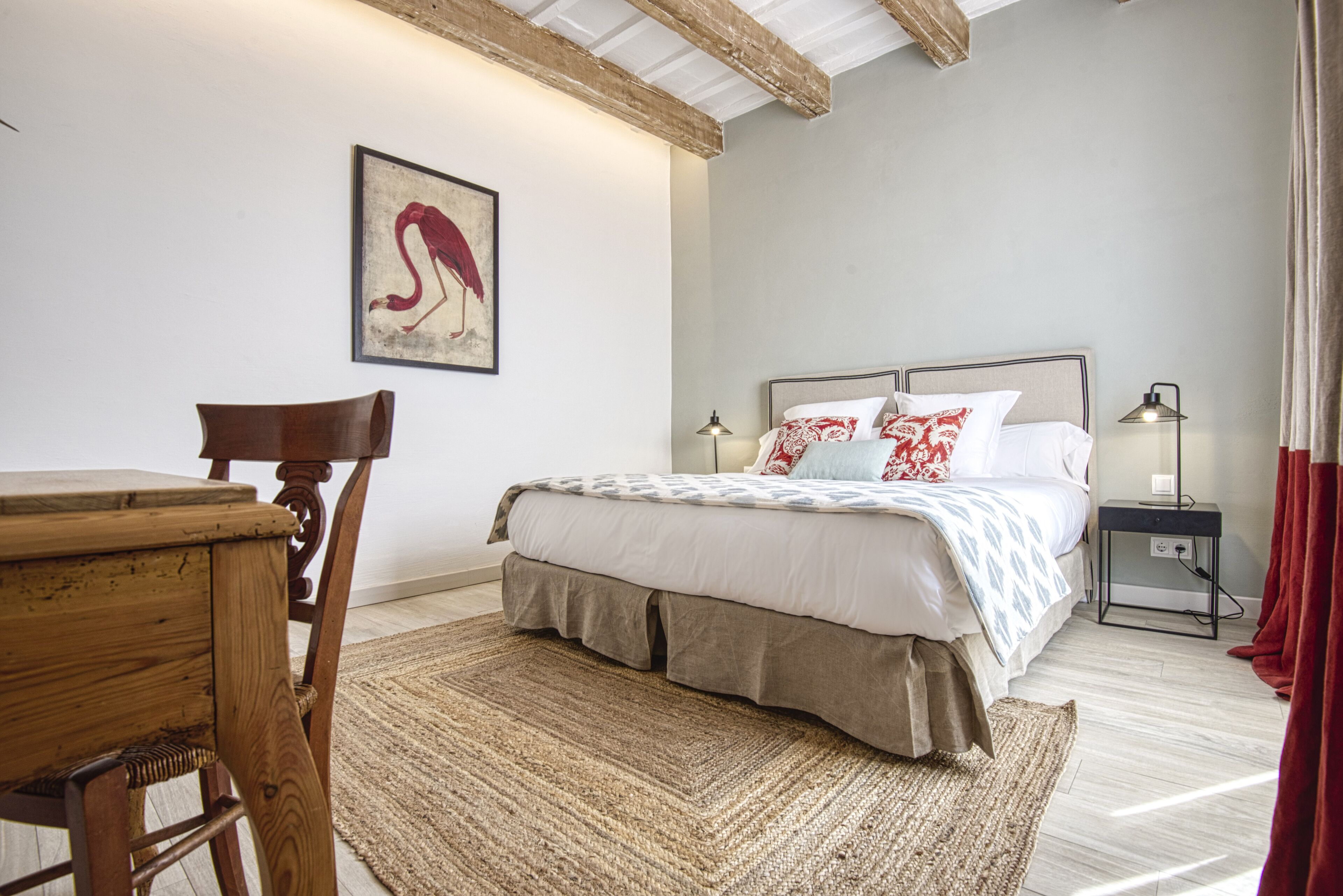 Een slaapkamer met een bed, een houten stoel, een tafel, een lamp en een schilderij aan de muur.