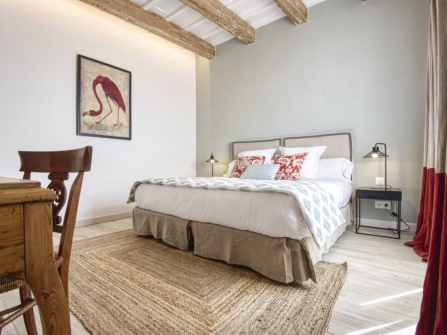 Een slaapkamer met een bed, een houten stoel, een tafel, een lamp en een schilderij aan de muur.