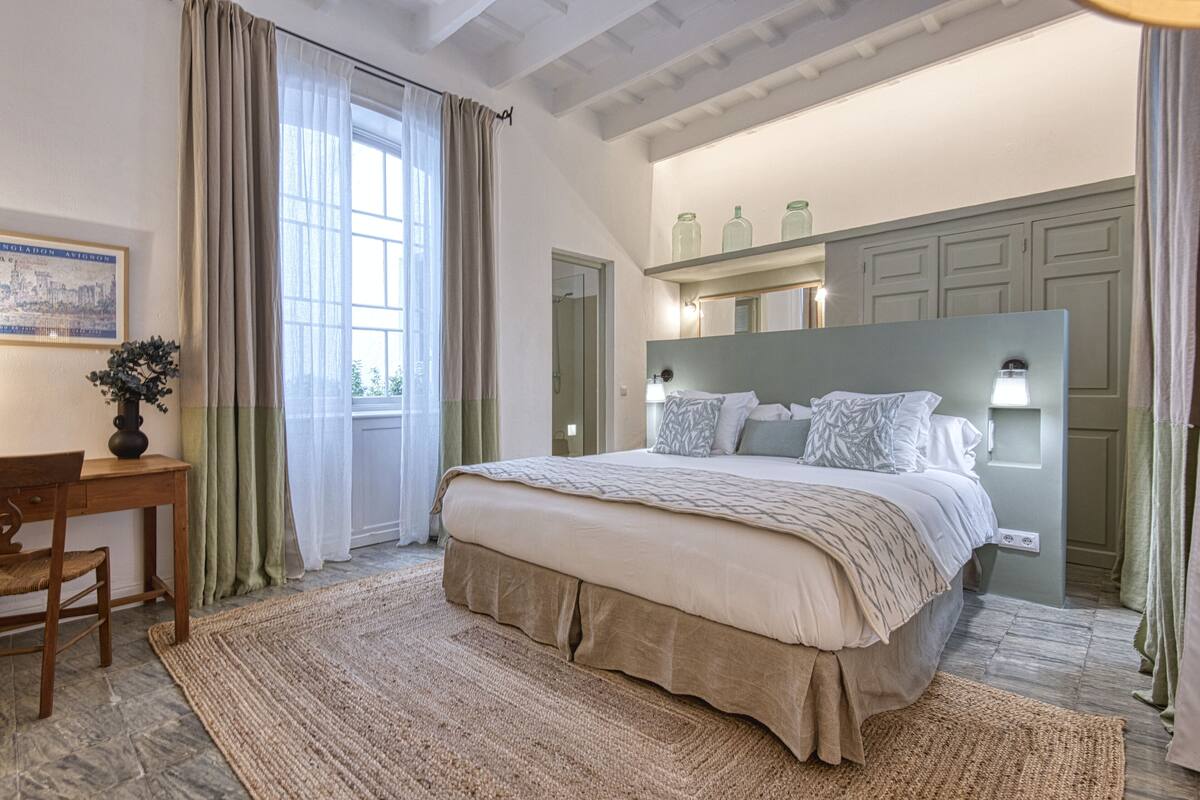 A bedroom with a large bed, a desk, and a chair. There is a window with curtains, a mirror, and a shelf with jars.