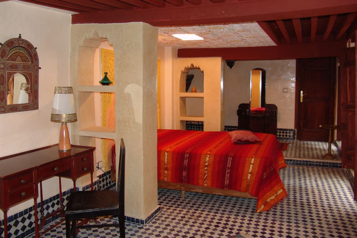 Une chambre d’hôtel avec un sol à carreaux, un lit recouvert d’une couverture rouge, un bureau avec une lampe et un miroir au mur.