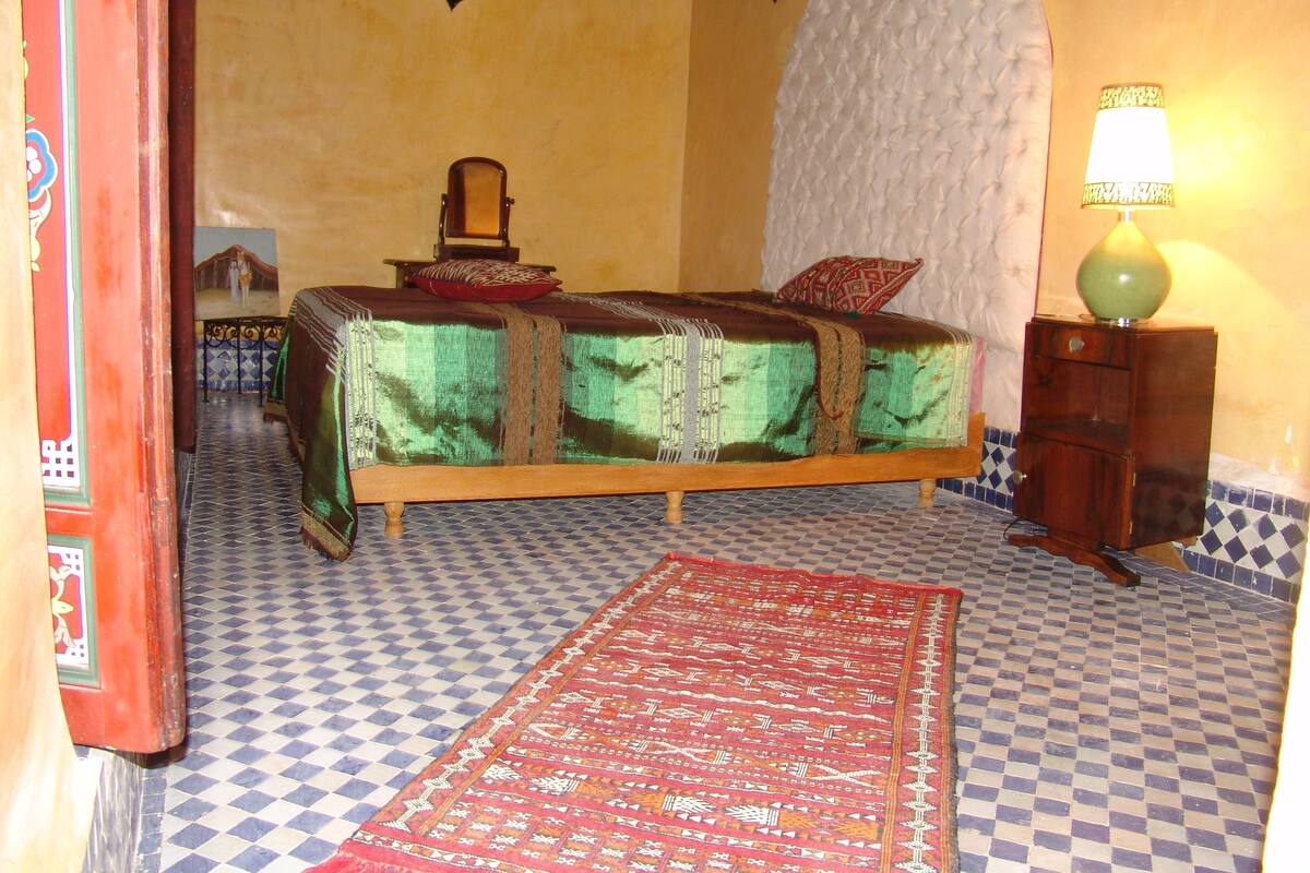 Une chambre avec un lit, une table de chevet, une lampe et un tapis à motifs.