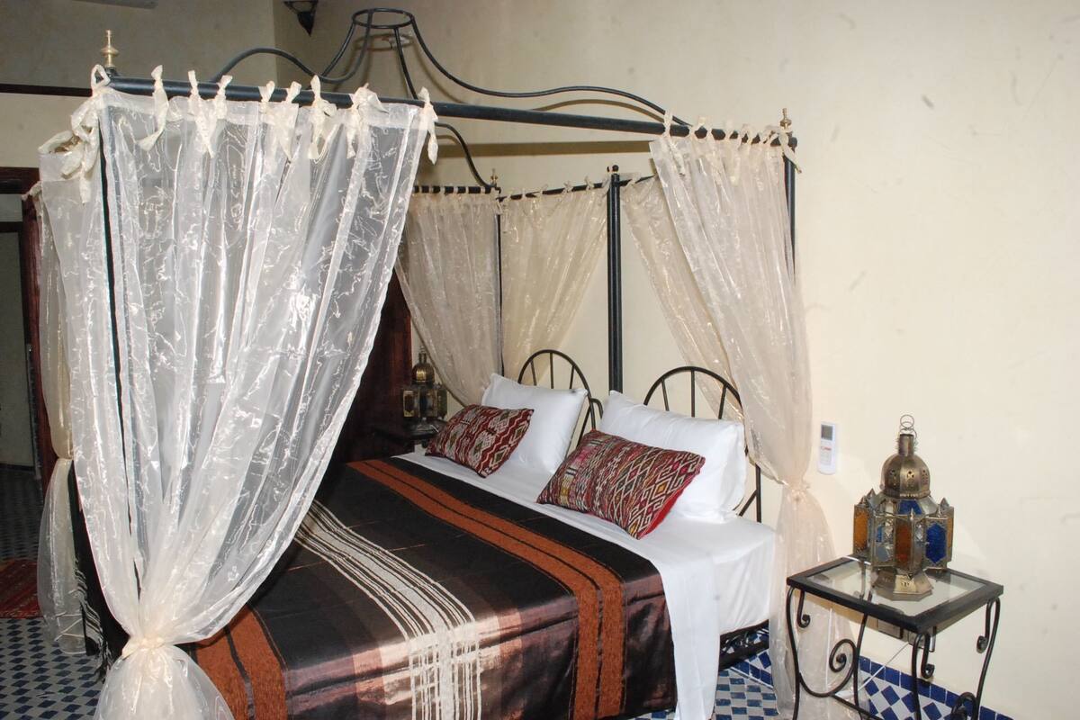 Un lit à baldaquin, deux oreillers, une table de chevet avec une bouteille, et un tapis à motifs.