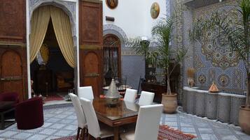 Un interior marroquí tradicional con una zona de comedor que incluye una mesa de madera y sillas blancas, rodeada de paredes con azulejos ornamentales y una alfombra estampada.