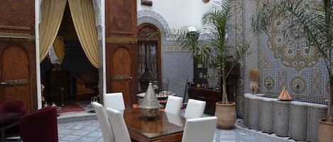 A traditional Moroccan interior with a dining area featuring a wooden table and white chairs, surrounded by ornate tile walls and a patterned rug.