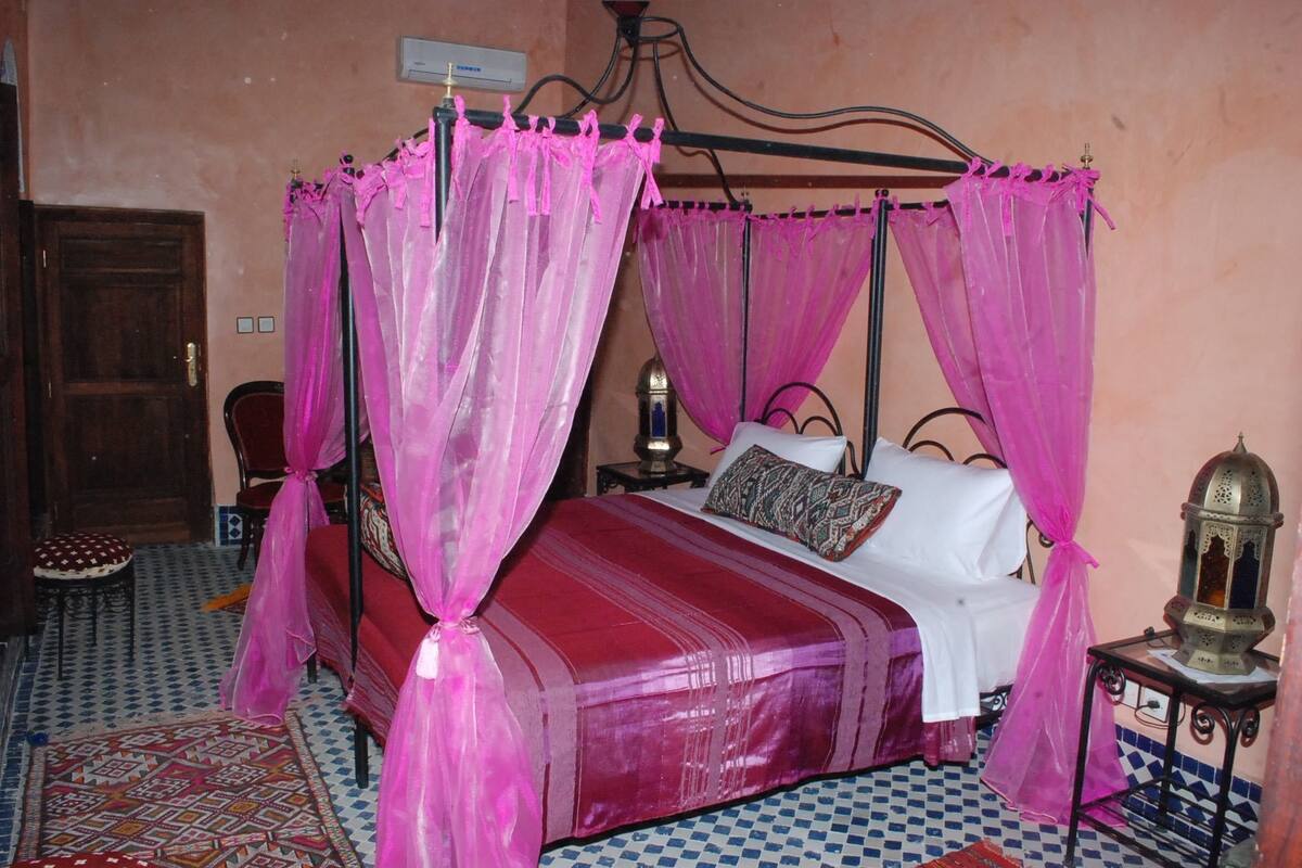 Un lit à baldaquin avec des rideaux roses, un tapis à motifs et une table de chevet avec une lampe.