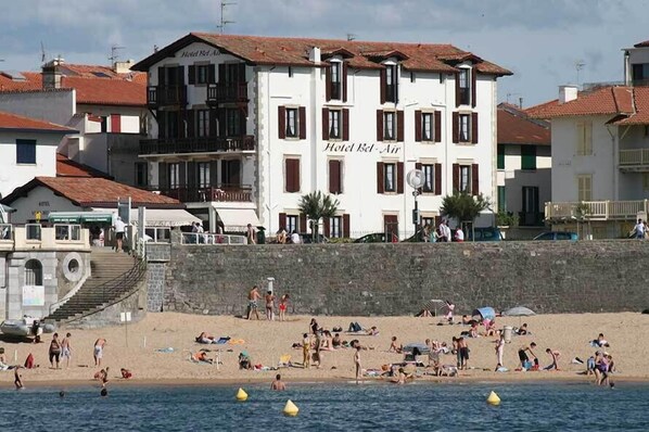 Un hôtel à la façade blanche et au toit recouvert de tuiles rouges, situé près d’une plage où les gens se prélassent au soleil et se baignent.