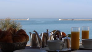 Een ontbijttafel met croissants, koffiepotten en glazen sinaasappelsap, met uitzicht op een kalme zee en een vuurtoren.