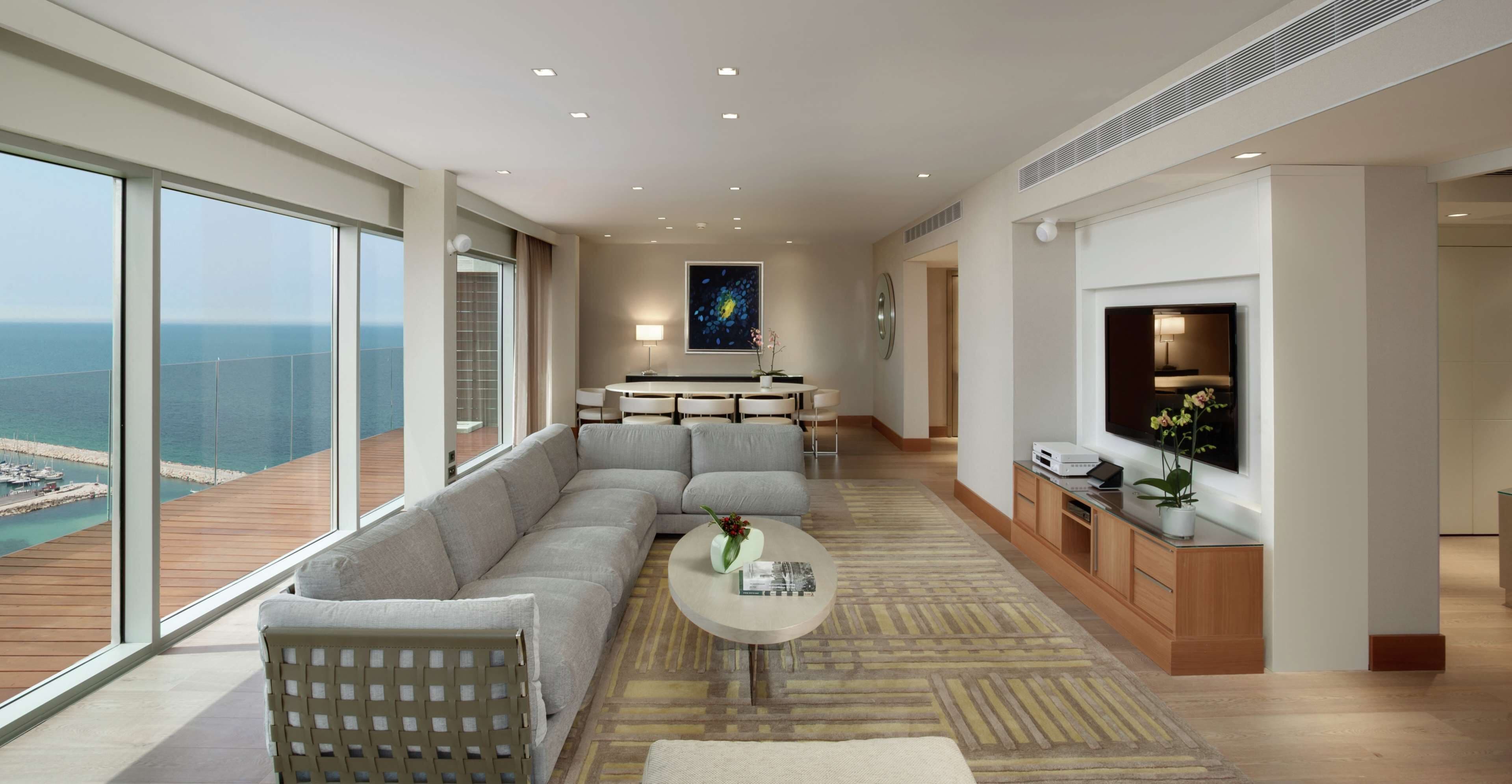 A modern living room with a large sectional sofa, a round coffee table, and a television. The room has large windows offering a view of the ocean and a dock.