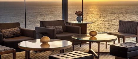 A modern lounge area with sofas, round tables, and a view of the ocean at sunset.