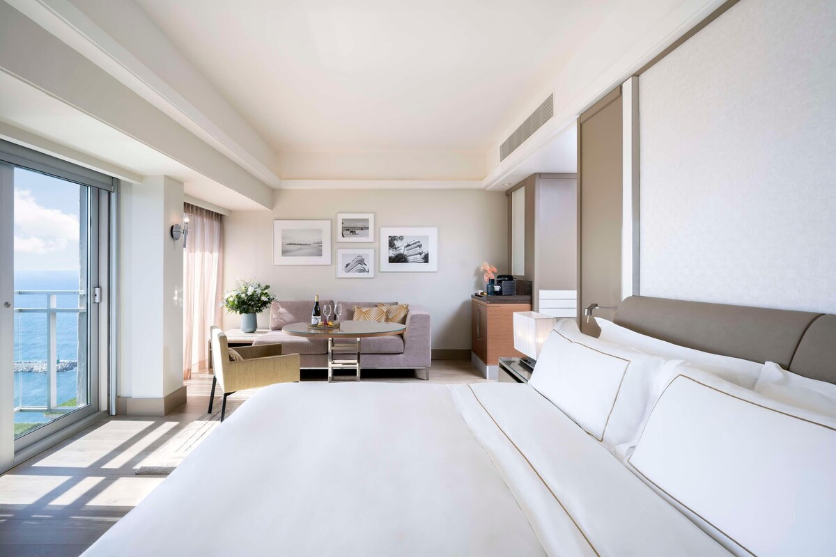 A modern hotel room with a large bed, a sofa, a dining table, and a view of the sea.