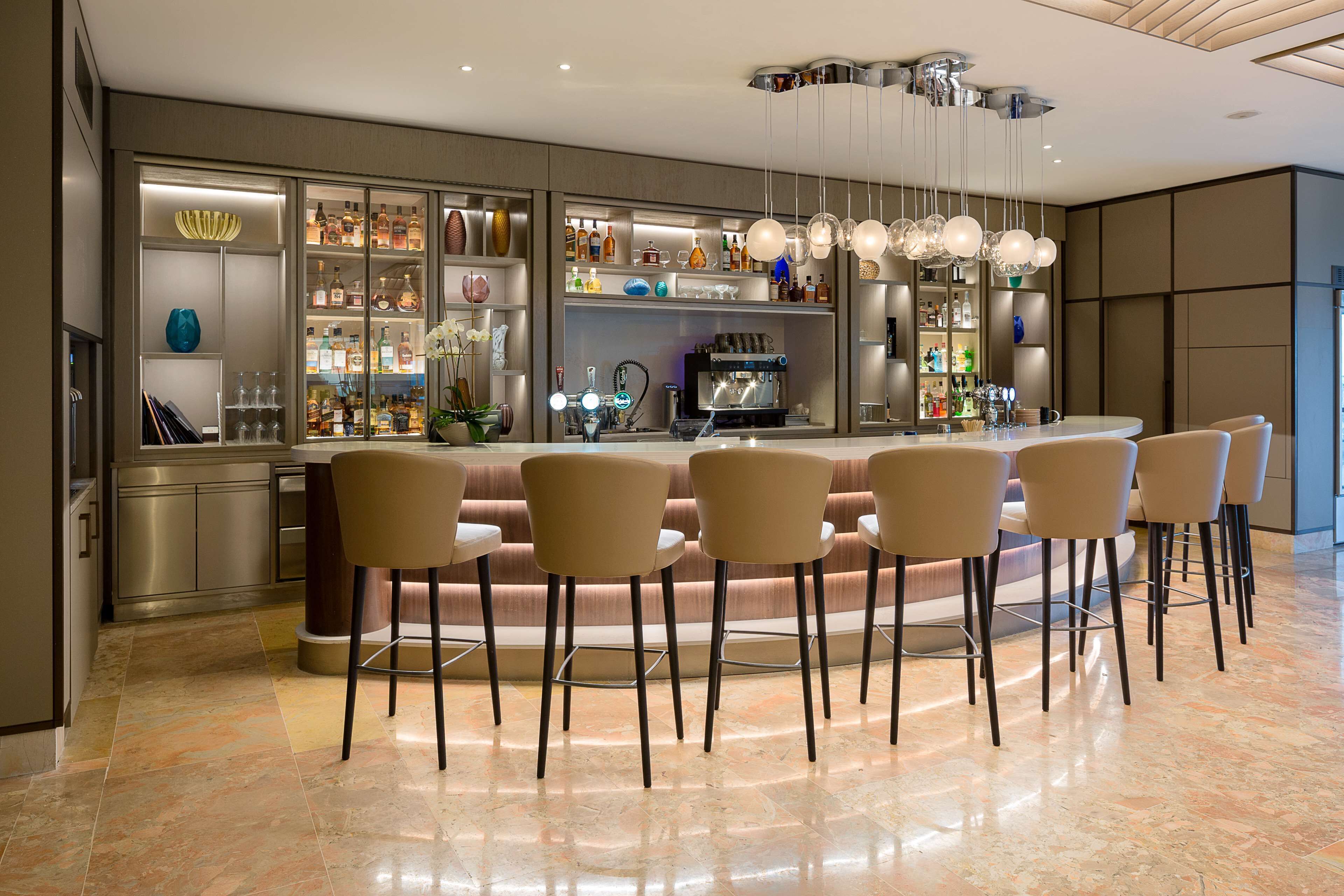 Un bar moderno con barra curva, diversas opciones de asientos y una amplia variedad de botellas y cristalería a la vista.