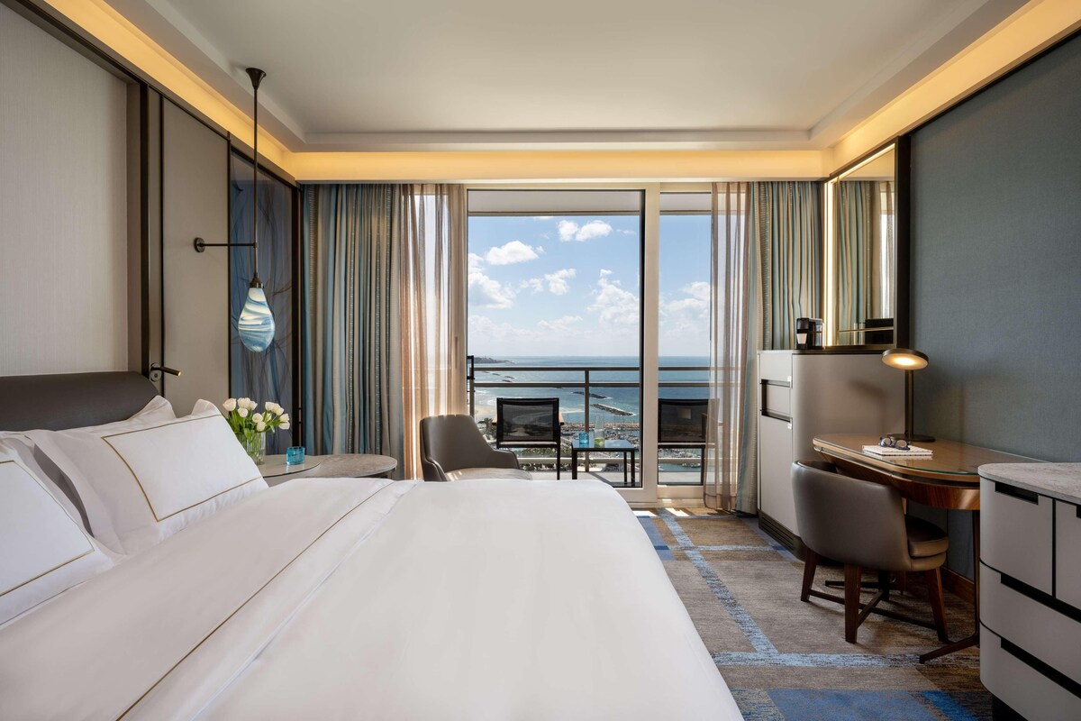 A hotel room with a large bed, a desk, a chair, and a view of the ocean.