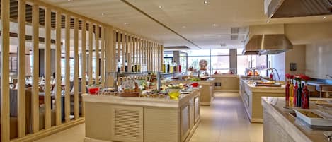 A modern hotel lobby with a central reception desk, a buffet area with various food items, and a seating area with tables and chairs.