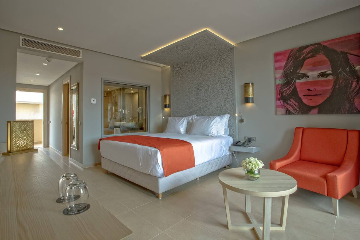 A modern hotel room with a bed, a red armchair, a small table with a vase, and a large abstract painting on the wall.
