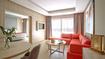 A modern hotel room with a sofa, a dining table with a vase of flowers, a TV, and a large window with curtains.