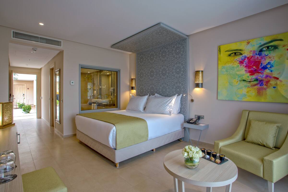 A modern hotel room with a large bed, a sitting area with a chair, a small table with a vase, and a painting on the wall.
