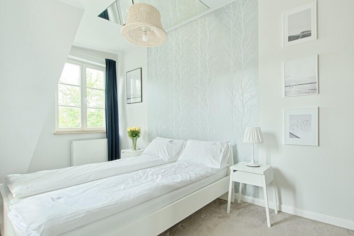 A bedroom with a bed, a bedside table with a lamp, a window with curtains, and wall-mounted pictures.