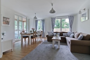 A modern living room with a sofa, dining table, chairs, and a coffee table. There are large windows with curtains, a ceiling light, and a framed picture on the wall. - Imperial Apartments - Valor (Sopot)